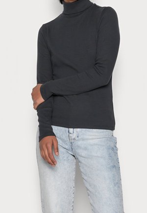 Personne portant un col roulé à manches longues gris foncé et un jean bleu clair délavé, avec un bras croisé sur l'autre.