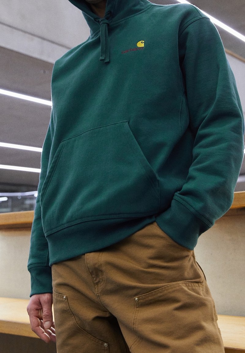 Mørkegrøn hoodie med en frontlomme, der har et lille gult og rødt logo. Sammen med brune bukser med synlige lommer og metaldetaljer.