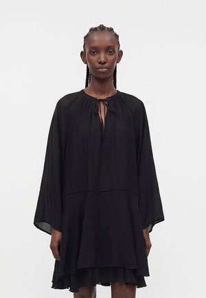 Femme aux cheveux tressés portant une robe noire ample avec de longues manches larges et une fermeture à nouer au niveau de l'encolure, debout devant un fond uni.