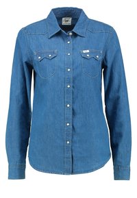 Chemise en denim bleu à manches longues avec boutons-pression à l'avant et deux poches poitrine, étiquetée avec l'étiquette de la marque Lee sur la poche droite.