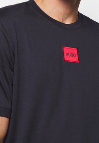 Námořnické bavlněné tričko s červenou vyšívanou nášivkou s textem "HUGO". Design zahrnuje kulatý výstřih a krátké rukávy.
