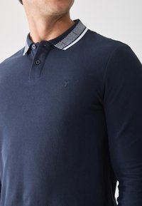 Marineblå langærmet polo-skjorte med en kontrasterende mønstret krave, to knapper og et diskret logo på brystet. Glat tekstur.