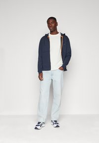 Marineblauwe zip-up hoodie met een oranje accent, gedragen over een licht t-shirt, gecombineerd met lichtblauwe jeans en witte sneakers. Eenvoudig ontwerp.