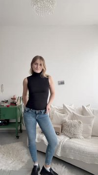 Top preto sem mangas com gola alta combinado com jeans skinny azul claro, usado com sapatos pretos. O fundo apresenta um sofá e itens de decoração.