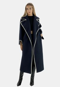 Langer marineblauer Mantel mit weißem Besatz, offenem Vorderteil und weitem Kragen. Hergestellt aus strukturiertem Stoff, mit Seitentaschen und kombiniert mit schwarzer Kleidung.