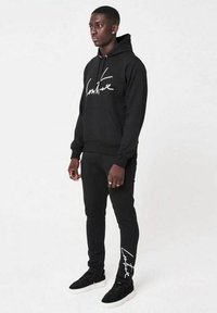 Svart hoodie och matchande byxor tillverkade av mjukt tyg. Hoodien har vit skrivstilstext på framsidan. Sneakers har en texturerad design.