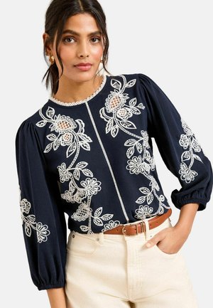 Femeie purtând o bluză bleumarin cu broderie florală albă și mâneci bufante, asortată cu pantaloni crem și curea maro.