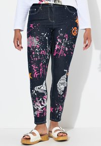 Vaqueros de mezclilla oscura con patrones de salpicaduras en rosa y blanco, gráficos de animales y acentos naranjas. Presentan un corte ajustado y un diseño tradicional de cinco bolsillos.