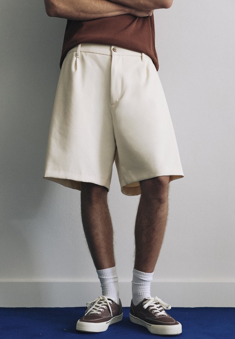 Personne portant un short crème jusqu’aux genoux, une chemise marron, des chaussettes blanches et des baskets marron, debout contre un mur uni, les bras croisés.