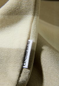 Gestrickter Stoff in Beige mit dezenten Strukturvariationen; enthält ein weißes Etikett mit dem Schriftzug "LINDBERGH" in schwarzer Schrift am Rand.