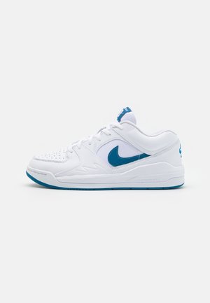 Hvid Nike lavt snørestøvle med blå swoosh, perforeret tå og blå ydersål, vist fra siden på en ensfarvet baggrund.