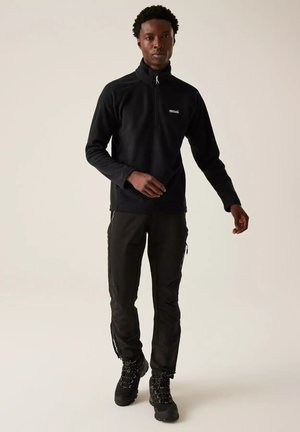 Hombre vestido con chaqueta de forro polar negra con media cremallera, pantalones cargo negros y botas de senderismo negras, de pie frente a un fondo claro y liso.