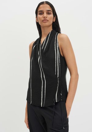 Femme portant un haut noir sans manches avec des rayures blanches verticales et un pantalon noir, debout avec une main dans la poche sur un fond uni.