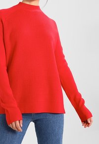 Red ribbed sweater with a high neck and long sleeves, featuring a relaxed fit and textured fabric. Paired with blue jeans.