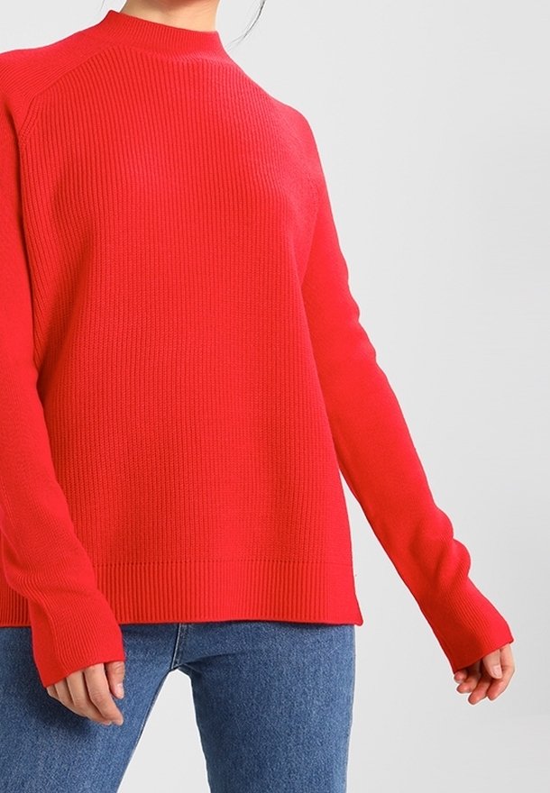 Red ribbed sweater with a high neck and long sleeves, featuring a relaxed fit and textured fabric. Paired with blue jeans.