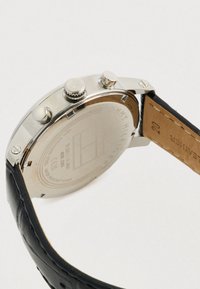 Ρολόι Tommy Hilfiger με ασημί θήκη, μαύρο δερμάτινο λουράκι και τρία κουμπιά στο πλάι σε λευκό φόντο.