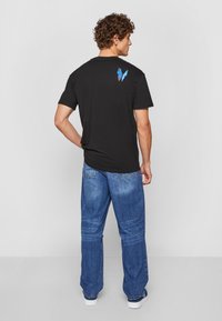 Sort bomuld T-shirt med et blåt sommerfugletryk på den øverste del af ryggen, parret med løstsiddende blå denimbukser og hvide sneakers.