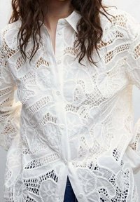 White embroidered blouse featuring intricate lace patterns, a button-up front, and a classic collar. Long sleeves with textured detailing.
