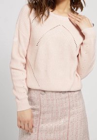 Pull rose clair en maille avec un motif texturé, présentant un motif ajouré. Porté avec une jupe ajustée rose et argentée à motif, ornée de boutons.