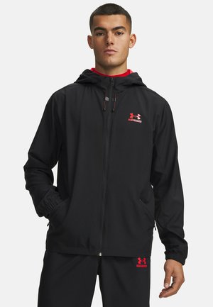 Mann trägt eine schwarze Under Armour Kapuzenjacke und passende Hose mit roten Logos, steht mit den Händen in den Jackentaschen.