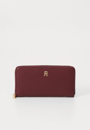 Burgundy wallet made of smooth leather, features a gold logo on the front, zip closure, and a compact rectangular shape.