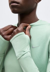 Κοντινό πλάνο ενός ατόμου που ρυθμίζει τον γιακά ενός μακρυμάνικου ανοιχτοπράσινου πουκαμίσου Nike με τρύπες για τους αντίχειρες στα μανίκια.