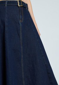 Falda de mezclilla azul oscuro con cintura ajustada y cinturón, bolsillos laterales y costuras amarillas en contraste. Diseño en línea A con textura suave.