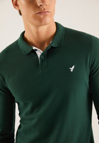 Mørkegrøn polo med lange ærmer, hvide detaljer på kraven og en lille hvid fuglelogo på venstre bryst. Materialet ser tekstureret ud.