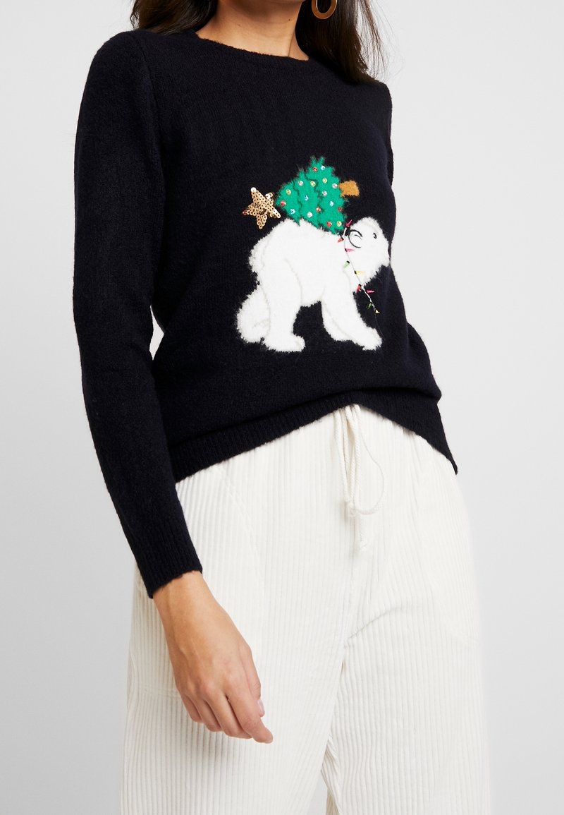 Donna che indossa un maglione nero con un orso bianco che porta un albero di Natale decorato di verde, abbinato a pantaloni bianchi a coste con coulisse.