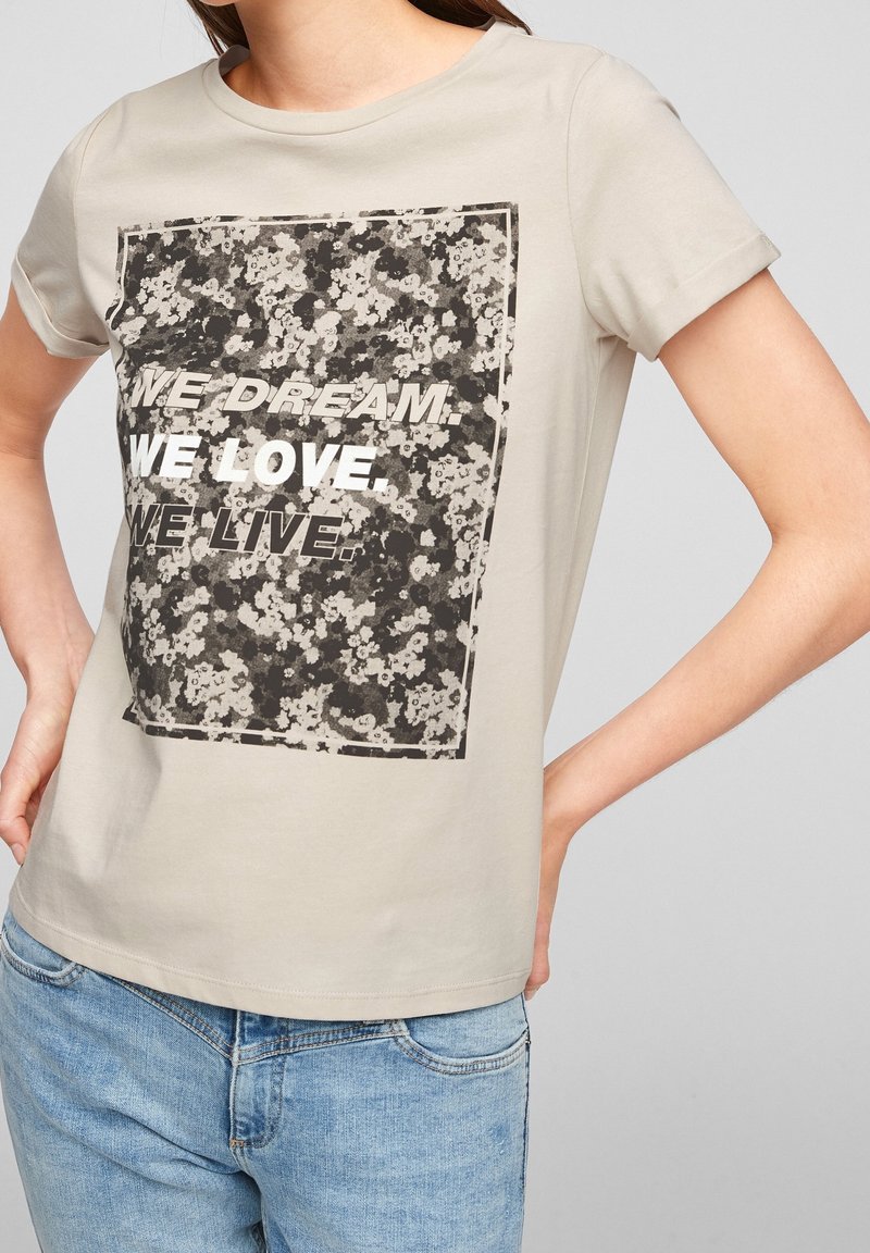 béžové tričko s ohrnutými rukávy, květinová čtvercová grafika a text "SNÍME. MILUJEME. ŽIJEME." kombinované se světle modrými džíny.