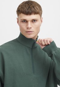 Sudadera verde con media cremallera que cuenta con un cuello alto, puños acanalados y un detalle del logo en el pecho. Textura suave, corte casual.