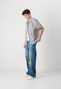Kortermet stripete skjorte i lys blå og hvit, sammen med løse, slitte blå jeans og hvite sneakers, stående mot en ensfarget bakgrunn.