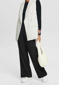 Weißer doppelt-knöpfiger Weste, schwarzes geripptes Langarmoberteil, schwarze weite Hose, weiße Sneaker, hält eine weiße Pufftasche.