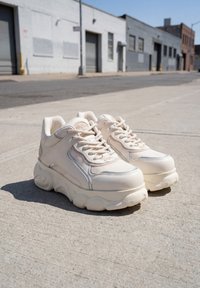 Ζευγάρι chunky sneakers εκρού με χοντρές σόλες τοποθετημένα σε πεζοδρόμιο από σκυρόδεμα σε αστικό χώρο με βιομηχανικά κτίρια.
