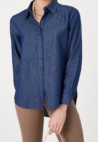 Mörkblå denimskjorta med knappstängning, spetsig krage, långa ärmar och en lätt rundad fåll. Slät struktur, visad på en modell.