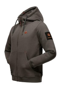 Harmaa vetoketjuhuppari, jossa on kiristettävä huppu, kaksi etutaskua, oranssit yksityiskohdat ja nappi vasemmassa hihassa. Pehmeä kankaan rakenne.