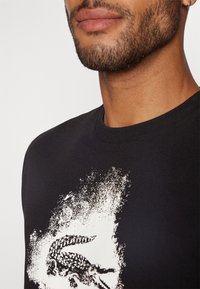 Mies, który ma na sobie czarną koszulę z białym graficznym nadrukiem krokodyla na klatce piersiowej, widoczna częściowo twarz i szyja.