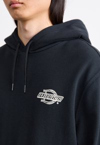 Sort bomulds-hoodie med en frontlomme og hætte med snøre. Har et hvidt "Dickies" logo på brystet. Glat tekstur.
