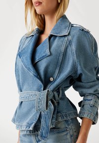 Giacca di denim in blu chiaro con vita cinturata, grande colletto e dettagli con bottoni. Presenta una vestibilità rilassata e un tessuto texturizzato.