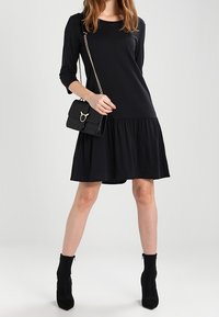 Robe noire à manches longues avec un bas évasé, en tissu doux ; associée à un petit sac à main noir avec une bandoulière chaîne.