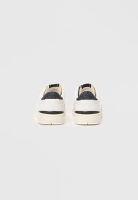 Baskets blanches avec un accent texturé gris au niveau du talon, une surface lisse et une semelle rembourrée. Présente une languette noire à l'arrière.