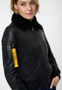 Black leather bomber jacket with a plush black collar, ribbed cuffs, zippered pocket on the sleeve, and a yellow branded tag attached.