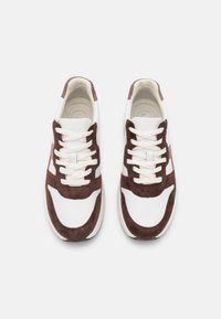 Białe i brązowe sneakersy z zamszowymi akcentami i tekstylną cholewką. Posiadają okrągłe sznurowadła, wyściełaną wkładkę i gumową podeszwę. Minimalistyczny design.