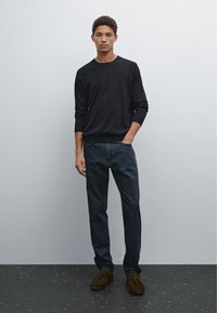 Mörk tröja med rund halsringning, kombinerad med mörka denimjeans och bruna mockaskor. Modellen står avslappnat med ena handen i fickan.