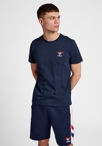 Marinblå bomulls-T-shirt med rund hals, prydd med en liten röd logotyp. Bärs tillsammans med matchande marinblå shorts som visar ett chevronmönster i rött och vitt.
