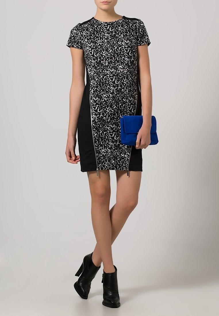 Femme portant une robe à manches courtes noire et blanche à motifs, des bottines noires, tenant une pochette bleue, debout devant un fond uni.