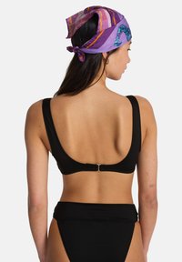 Vrouw van achteren gezien, gekleed in een zwarte bikini en een paars sjaal met patroon, vastgebonden in de nek, met een kleine tatoeage op de arm zichtbaar.