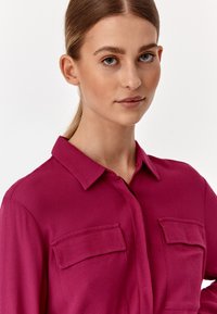 Fuksia siidist särk kraega, millel on kaks rinna taskut ja sile tekstuur. Nööbitav eesmine osa.