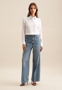 Chemise blanche en lin à boutons associée à un jean large bleu clair et des sandales à talons noires, avec une coupe décontractée et un design épuré.