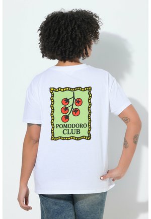 Personne aux cheveux bouclés portant un t-shirt blanc avec une branche de tomate rouge et le texte « POMODORO CLUB » dans le dos, debout contre un mur blanc.